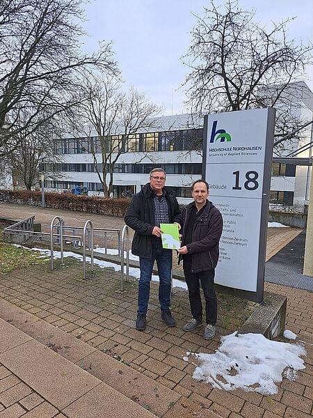 Heiko Lange und Prof. Dr. Mario Schölzel stehen nebeneinander vor der Hinweistafel von Haus 18. Sie halten gemeinsam eine grüne HSN-Dokumentenmappe. Im Hintergrund Haus 18. Auf dem Boden liegen Schneereste, die Bäume sind kahl.