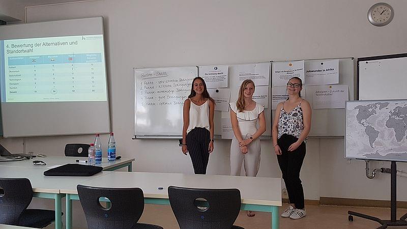 Drei junge Menschen stehen lächelnd nebeneinander. Im Hintergrund eine Präsentation mittels Beamer und Whiteboard.