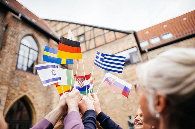 Eine Gruppe von Menschen hält kleine Flaggen verschiedener Länder zusammen nach oben. Im Hintergrund ist ein historisches Gebäude zu erkennen.