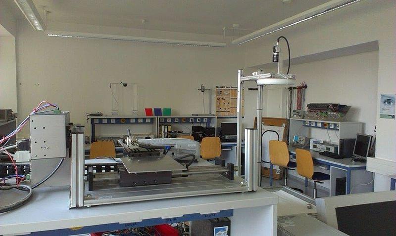 Ein Laborraum mit mehreren technischen Apparaturen und Computern. Im Vordergrund befindet sich ein großer Aufbau aus Metallprofilen, der für bildverarbeitende Experimente genutzt wird. Im Hintergrund sind mehrere Arbeitsplätze mit Computern und Laborgeräten zu sehen. Die Arbeitsplätze sind mit Stühlen ausgestattet, und verschiedene elektronische Geräte und Werkzeuge sind auf den Tischen und Regalen verteilt. Die Laborumgebung ist sauber und gut organisiert.