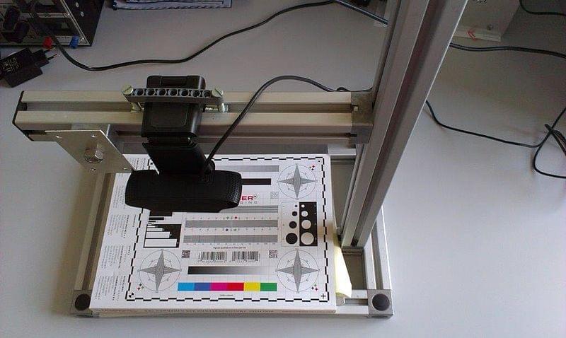 Eine technische Einrichtung in einem Labor zur Bildverarbeitung. Auf einem Tisch steht ein Rahmen aus Metallprofilen mit einer befestigten Kamera, die auf ein Testmuster mit verschiedenen Farben, Graustufen und Markierungen gerichtet ist. Das Testmuster liegt auf einer weißen Unterlage, und Kabel verbinden die Kamera mit anderen Geräten außerhalb des Bildes.