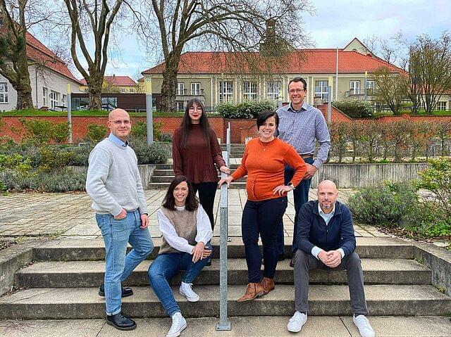 Eine Gruppe von sechs Personen posiert lächelnd auf einer Treppe im Freien. Im Hintergrund sind Gebäude mit roten Dächern und Bäume ohne Blätter zu sehen. Die Personen sind unterschiedlich gekleidet: zwei Männer in Pullovern, eine Frau in einem roten Pullover, eine Frau in einem braunen Pullover, ein Mann in einem karierten Hemd und ein Mann in einer blauen Jacke. Die Szene wirkt entspannt und freundschaftlich.