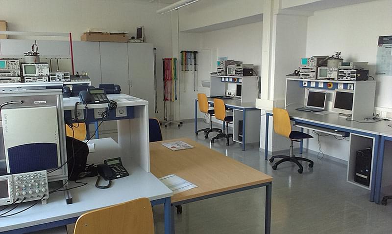Das Bild zeigt mehrere Arbeitsplätze mit Schreibtischen und Stühlen. Auf den Schreibtischen stehen verschiedene technische Geräte, darunter Oszilloskope, Spektrumanalysatoren, Netzgeräte und Computer. Im Hintergrund sind Schränke und Regale mit weiterem Laborequipment zu sehen. Die Wände des Raumes sind weiß gestrichen, und die Fenster lassen Tageslicht herein, das den Raum erhellt.