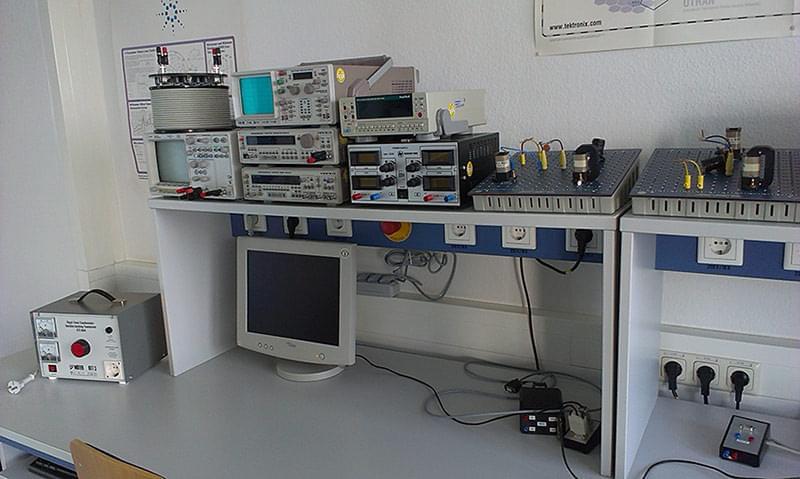 Ein Kommunikationslabor mit verschiedenen technischen Geräten und Instrumenten. Im Vordergrund steht ein Computermonitor, dahinter sind mehrere Messgeräte, Oszilloskope und Netzteile aufgestapelt. Rechts davon befinden sich weitere Geräte und Module, die auf einem Tisch angeordnet sind. Die Wände des Raums sind mit verschiedenen Plakaten und Diagrammen dekoriert. Das Labor ist hell und gut beleuchtet, mit einem großen Fenster im Hintergrund, das natürliches Licht hereinlässt. Es gibt mehrere Arbeitsstationen mit Computern und Stühlen.