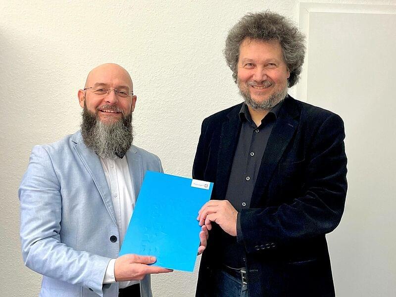 Ein Mann mit grauem Haar und Bart, gekleidet in einem dunklen Anzug, überreicht einem anderen Mann, ebenfalls mit Bart und in einem hellen Anzug, eine blaue Ernennungsurkunde. Beide Männer lächeln in die Kamera, während sie die Urkunde gemeinsam halten.