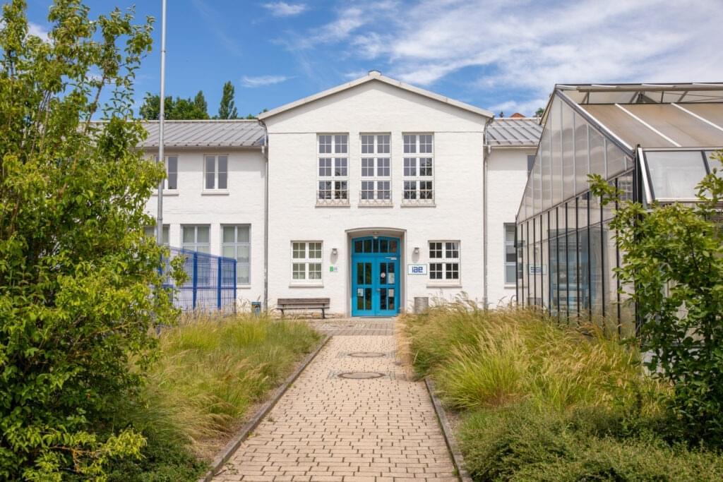 Haus 25 - IAE - Ein weißes Gebäude mit blauer Tür. Ein gepflasterter Weg führt drauf zu. Rechts ein Gewächshaus, Links ein Baum.