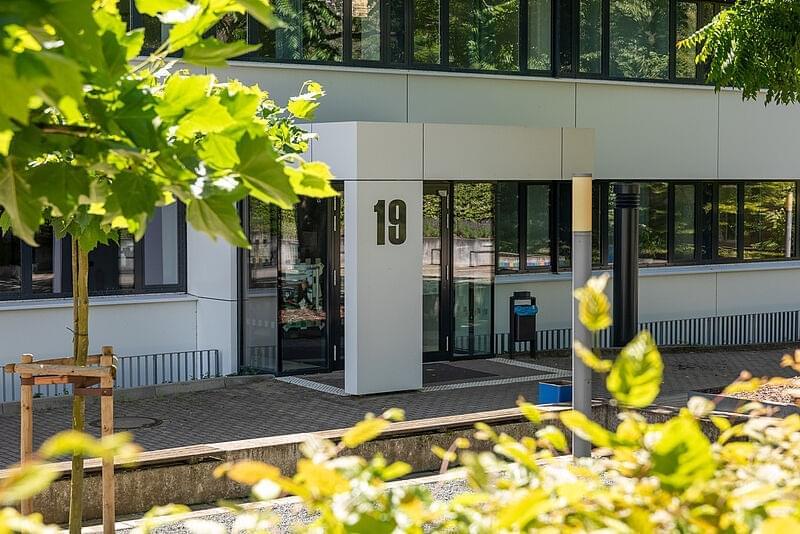 Eingangsbereich von Haus 19. Im Vordergrund Bäume mit grünem Laub.