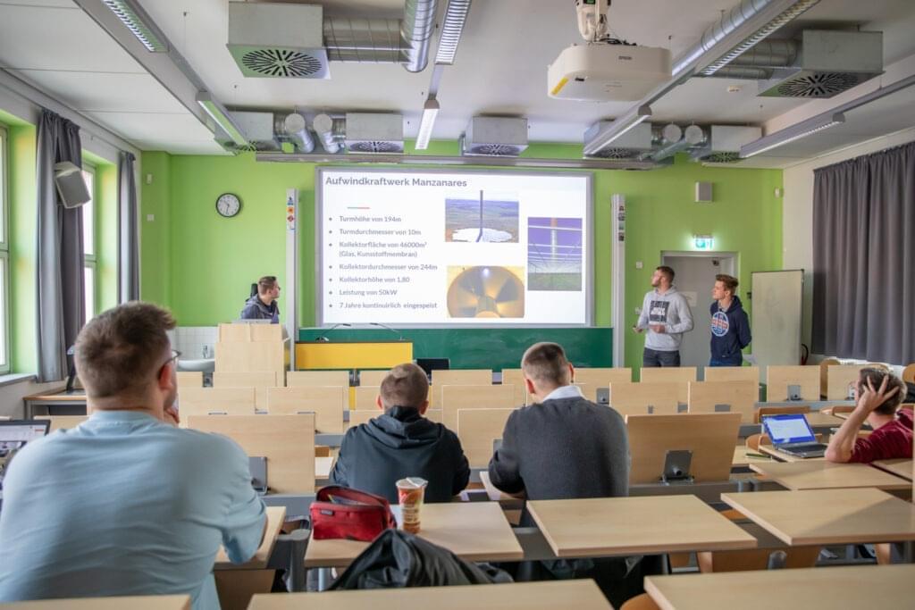 Im Hörsaal der Hochschule präsentieren zwei Studierende vor einer Projektionswand ein Thema über das Aufwindkraftwerk Manzanares. Die Präsentationsfolie enthält technische Daten wie die Turmhöhe von 194 m, den Turmdurchmesser von 10 m, und die Kollektorfläche von 46.000 m². Weitere Studierende sitzen im Hörsaal und folgen der Präsentation.