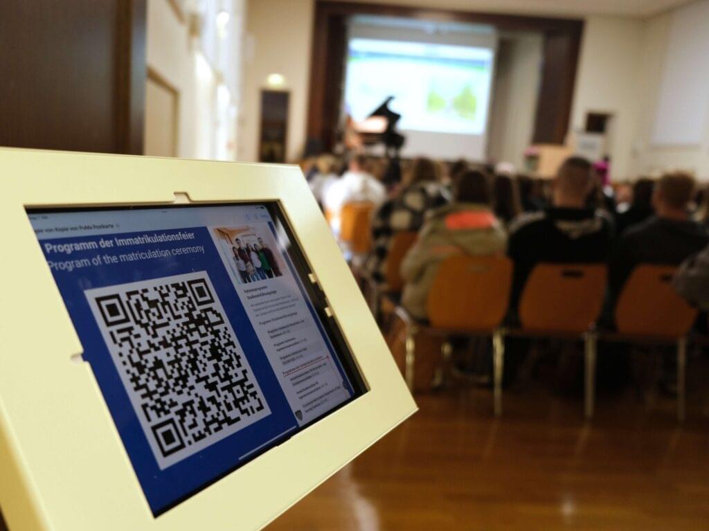 Tablet mit QR-Code zum Programm, im Hintergrund das Audimax mit Studierenden