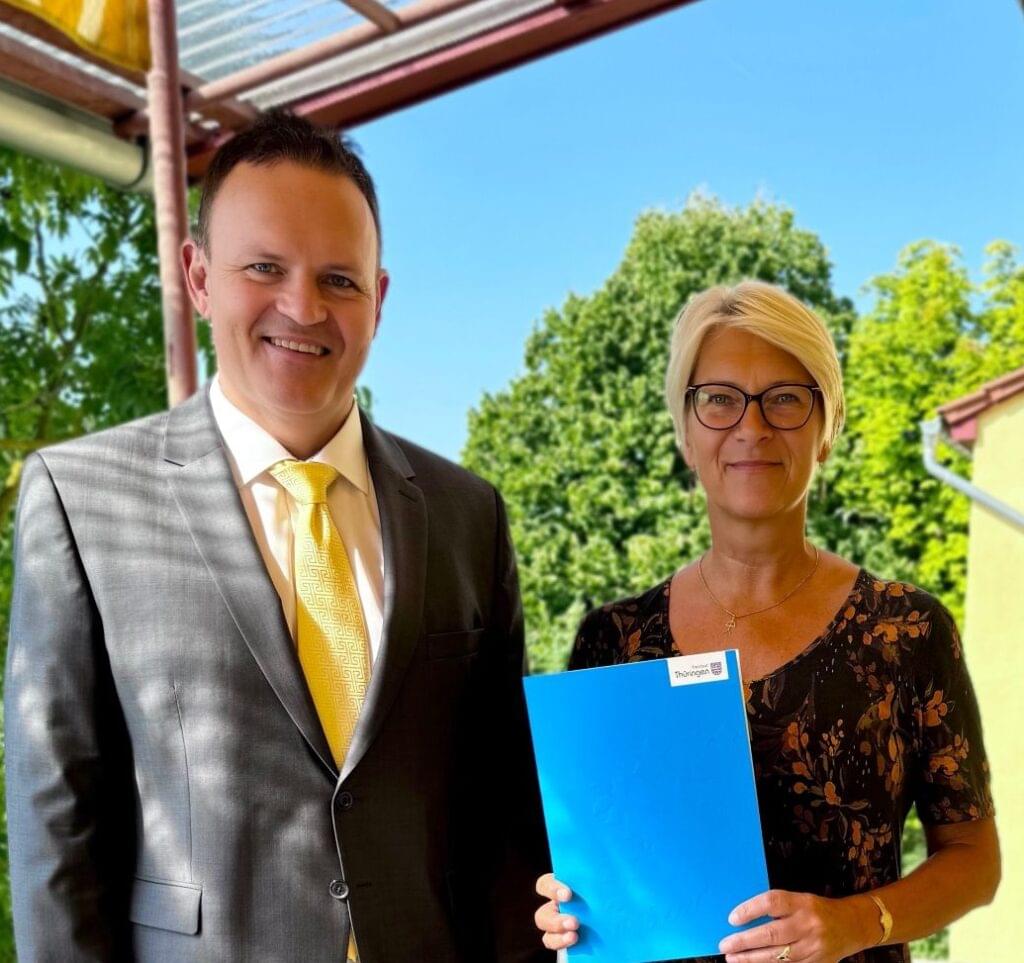 Das Bild zeigt zwei Personen, die nebeneinander auf einem sonnenbeschienenen Balkon oder einer Terrasse stehen und in die Kamera lächeln. Der Mann links trägt einen dunklen grauen Anzug mit einer gelben Krawatte, während die Frau rechts ein schwarzes, geblümtes Kleid trägt und eine blaue Mappe in den Händen hält. Im Hintergrund sind Grünpflanzen und ein Teil des Gebäudes zu sehen.