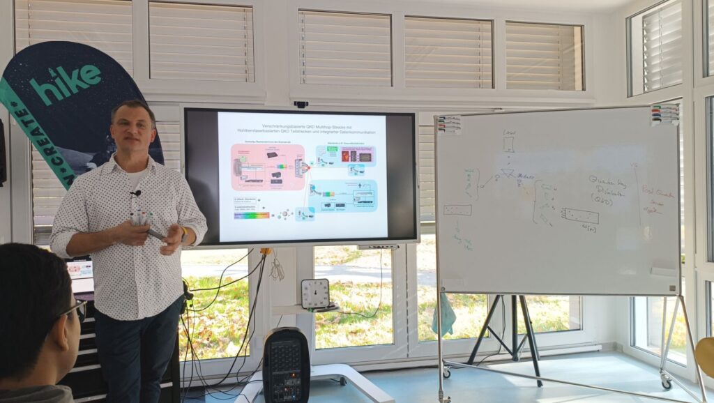 Prof. Hühn steht vor einem Bildschirm und einem Whiteboard in einem Seminarraum. Auf dem Bildschirm ist eine schematische Darstellung einer verschränkungsbasierten optischen Kommunikationsstrecke zu sehen. Er hält mehrere Stifte in der Hand und gibt eine Präsentation. Im Hintergrund sind Fenster und eine „hike“-Werbeflagge zu sehen.
