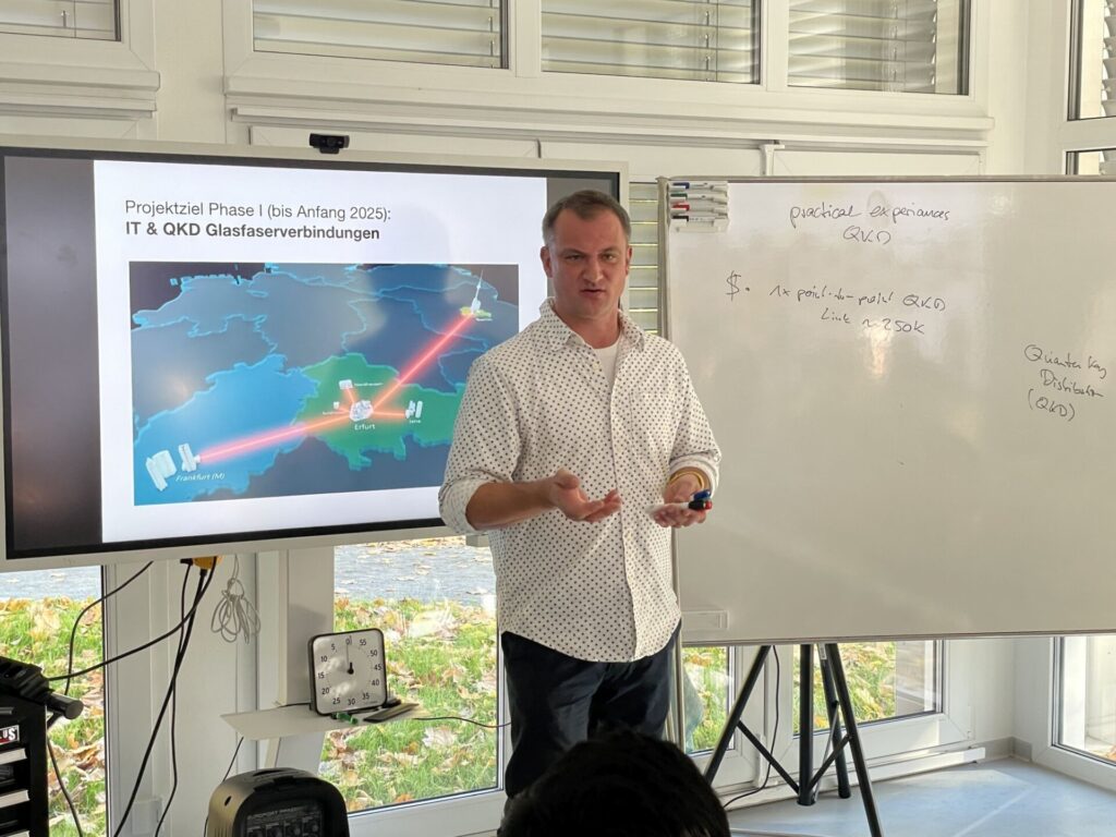 Prof. Hühn präsentiert vor einem Bildschirm und einem Whiteboard. Auf dem Bildschirm ist eine Karte mit dem Projektziel „IT & QKD Glasfaserverbindungen“ bis Anfang 2025 zu sehen, die verschiedene Städte wie Erfurt und Frankfurt zeigt. Der Mann hält einen Stift und erläutert das Thema der Quantenschlüsselverteilung.