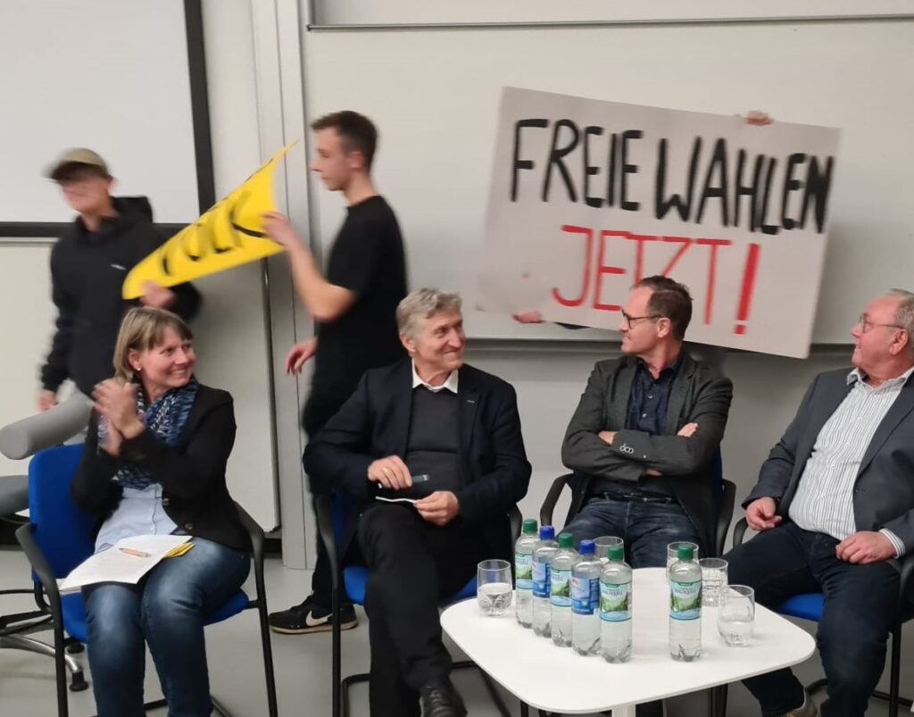 Podiumsdiskussion an der Hochschule Nordhausen: Zeitzeugen und Gäste diskutieren über die Bedeutung freier Wahlen, während engagierte Studierende die Demonstrationsstimmung von 1989 mit nachgestellten Transparenten lebendig werden lassen.