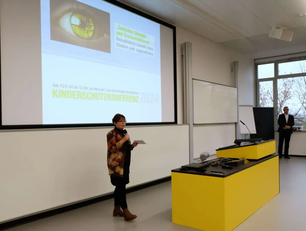 Eine Rednerin steht in einem Hörsaal vor einer Projektionsfläche und hält einen Vortrag bei der Kinderschutzkonferenz 2024 der Hochschule Nordhausen. Auf der Präsentation steht der Titel „Zwischen Neugier und Grenzverletzung – Sexualisierte Gewalt unter Kindern und Jugendlichen“. Der Vortragssaal ist hell und modern gestaltet.