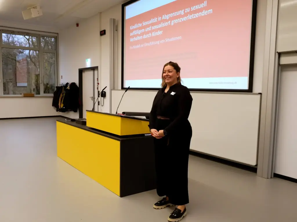 Eine Referentin steht vor einer Projektionsfläche mit dem Thema „Kindliche Sexualität in Abgrenzung zu sexuell auffälligem und sexualisiert grenzverletzendem Verhalten durch Kinder“. Sie lächelt.