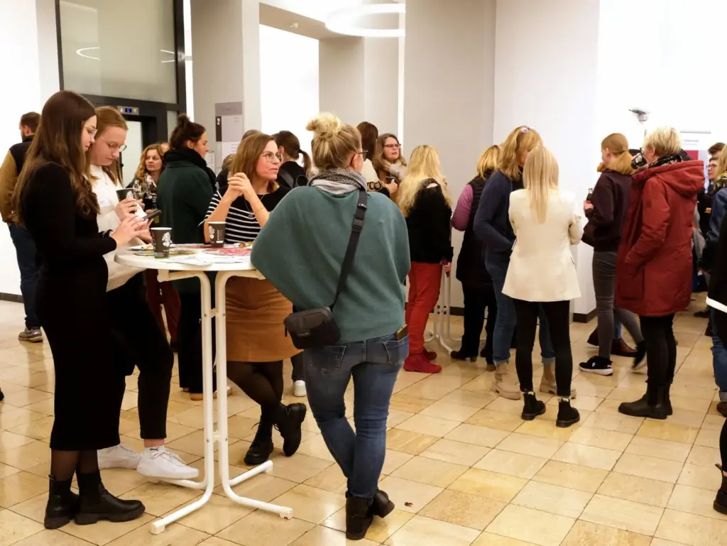 Eine Gruppe von Teilnehmenden steht in einem hellen Foyer bei einem Stehempfang. Einige Personen unterhalten sich in kleinen Gruppen, während andere Getränke und Snacks genießen.