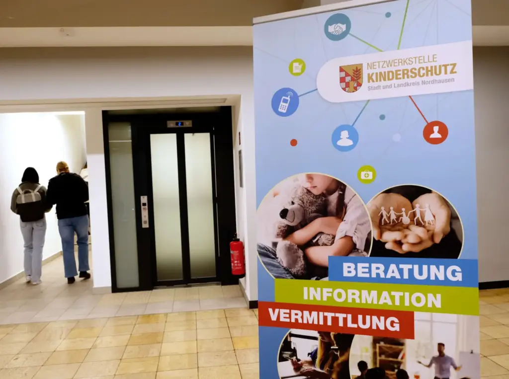 Ein Roll-Up mit der Aufschrift „Netzwerkstelle Kinderschutz – Stadt und Landkreis Nordhausen“ steht in einem Eingangsbereich. Das Banner hebt die Themen Beratung, Information und Vermittlung hervor und zeigt Bilder von Kindern und Familien. Im Hintergrund ist eine Tür zu sehen, durch die zwei Personen gehen.