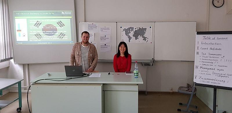 Zwei Personen stehen nebeneinander hinter einem Schreibtisch. Im Hintergrund eine Präsentation per Beamer und Whiteboards.