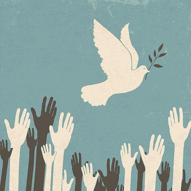 Eine Illustration zeigt eine weiße Taube, die einen Olivenzweig im Schnabel trägt und über mehrere ausgestreckte Hände fliegt. Die Hände sind in verschiedenen Hauttönen dargestellt und strecken sich nach der Taube. Der Hintergrund ist in einem blassen Blau gehalten, was der Szene eine friedliche Atmosphäre verleiht.