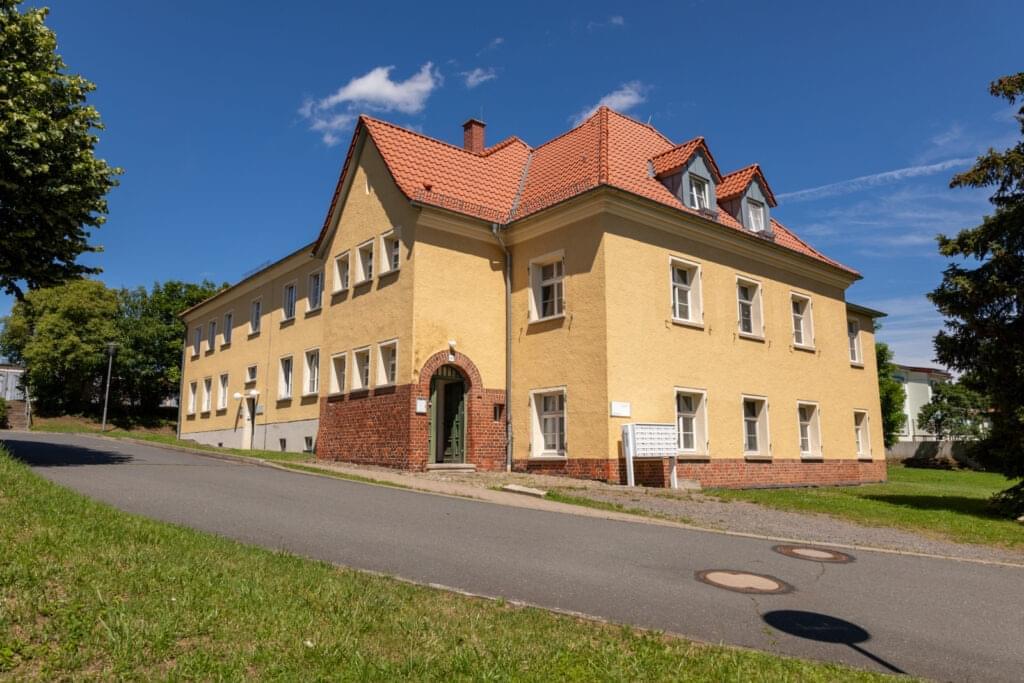 Ein älteres, gelbes Gebäude mit rotem Ziegeldach. Der Himmel ist blau. Davor führt eine schmale Straße entlang.