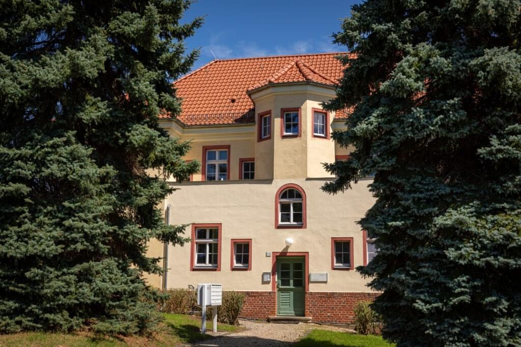 Ein älteres Gebäude mit einem roten Ziegeldach und einem kleinen Türmchen. Links und rechts stehen Nadelbäume.