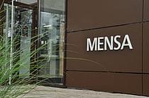 Rechts die braune Hauswand mit dem großen weißen Schriftzug "MENSA". Links davon die geöffnete Tür zur Mensa. Links im Vordergrund ragen Gräser ins Bild.